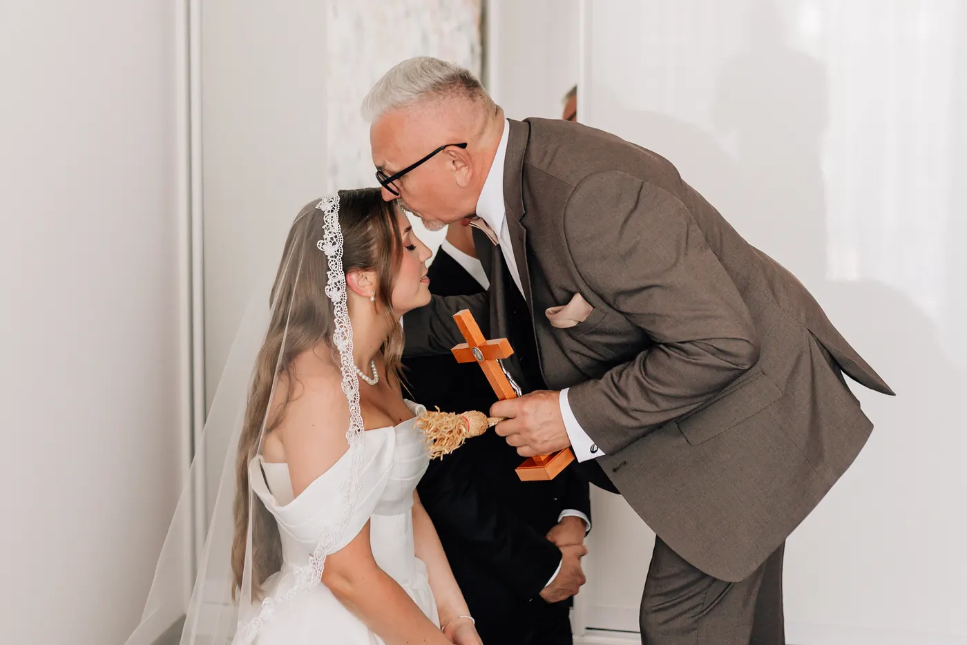 Tata z krzyżem w dłoni całuje pannę młodą w czoło podczas błogosławieństwa